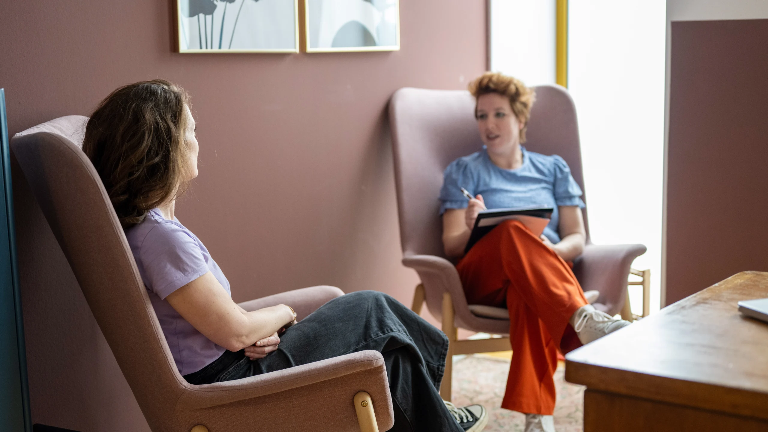 A therapy session is taking place in a warm and inviting room. Two women are seated in comfortable chairs, facing each other.